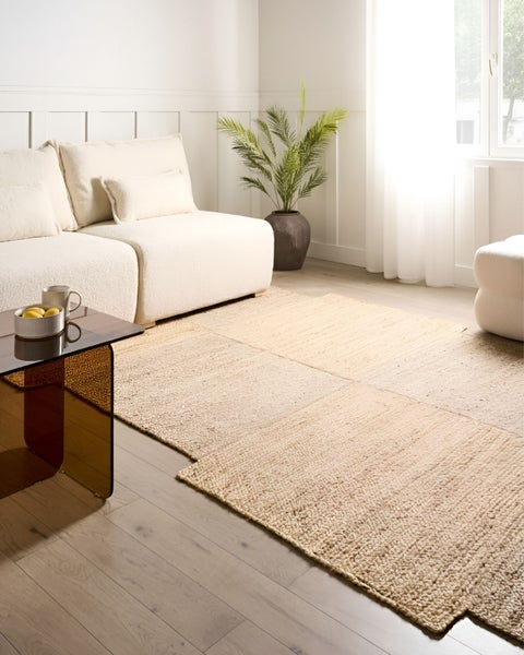 Gewebter Teppich aus Naturmaterial in einem hellen, modernen Wohnzimmer mit Sofa, Glastisch und Zimmerpflanze.
