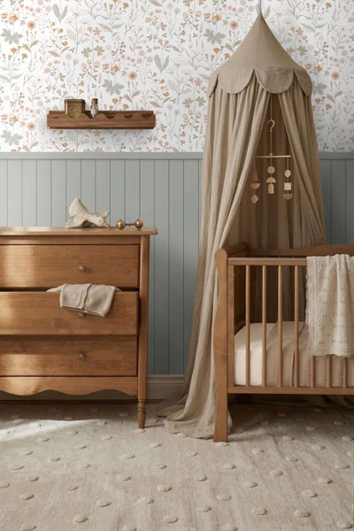 Ein gemütliches Babyzimmer mit Holzmöbeln, floraler Tapete, einem Kinderbett mit Betthimmel, einer Kommode und einem Wandregal.