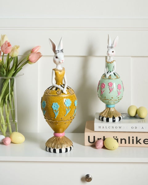 Dekorative Osterhasen-Figuren mit eiförmigen Kleidern und Blumenmustern, arrangiert mit Tulpen und Ostereiern in einer hellen, organisierten Szene.