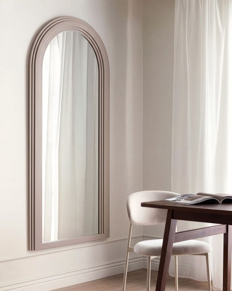 Wandspiegel mit gewölbtem Rahmen im modernen Stil hängend an einer Wand neben einem Holztisch mit Stuhl in einem hellen Raum.