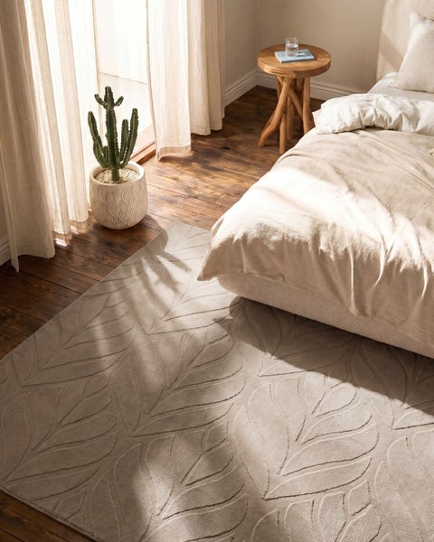 Hell eingerichtetes Schlafzimmer mit einem Teppich mit Blattmuster auf Holzboden neben einem Bett und einem Kaktus im Topf.