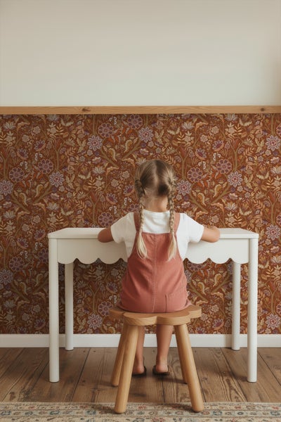 Kind sitzt an einem weißen Kinderschreibtisch vor einer gemusterten Tapete.