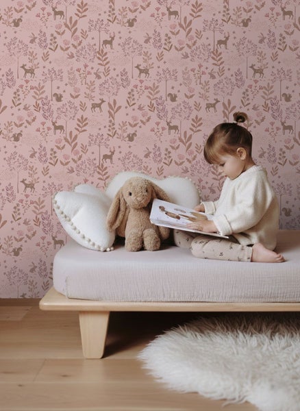 Ein Mädchen sitzt auf einem Kinderbett vor einer Tapete mit Waldmotiven, die Hirsche, Eichhörnchen und Pflanzen zeigt.
