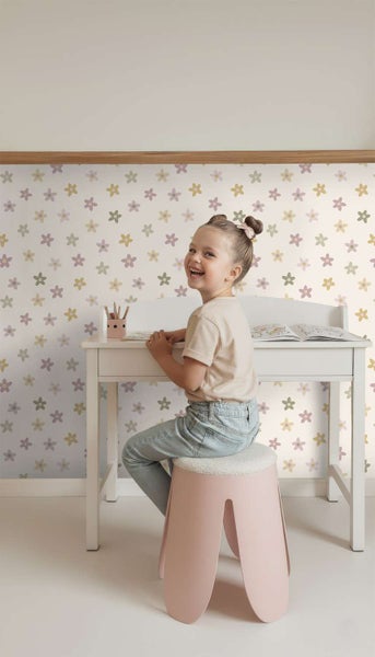 Ein lächelndes Kind sitzt auf einem Hocker an einem Kinderschreibtisch vor einer Wand mit Tapete mit Blumenmuster in einem Kinderzimmer.