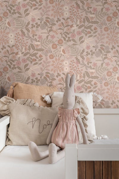 Vliestapete mit Blumen, Vögeln und Schmetterlingen in einem hellen Kinderzimmer hinter einem Bett mit Kissen und Stofftier.