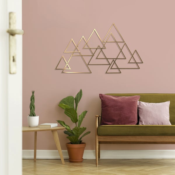 Wanddekoration aus Metall mit geometrischem Dreiecksmuster in einem hellen Wohnzimmer mit Sofa aus Holz und Zimmerpflanzen.