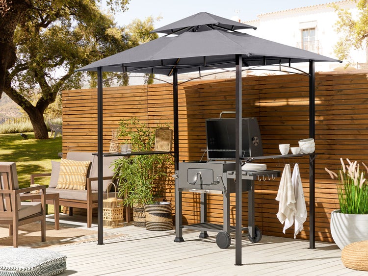 Moderner Grillpavillon mit Textildach über Holzkohlegrill auf einer hellen Terrasse mit Holzmöbeln, Pflanzen und Holzzaun.