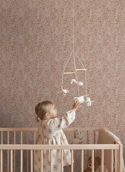 Mädchen im Kinderbett, das ein Mobile mit floral gemusterter Tapete im Hintergrund berührt