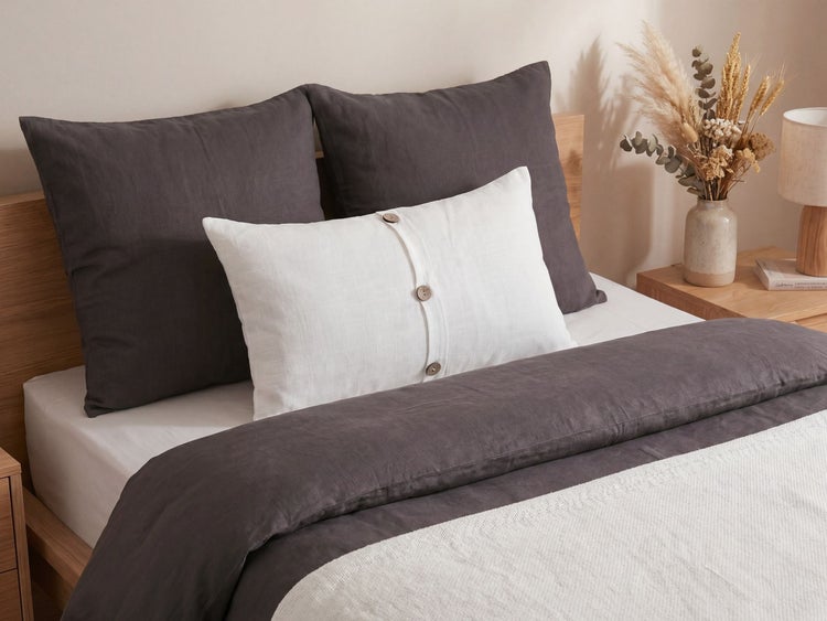 Helles Schlafzimmer mit Holzbett, strukturierter Bettwäsche und Zierkissen mit Knopfleiste, Nachttisch mit Vase und Trockenblumen.