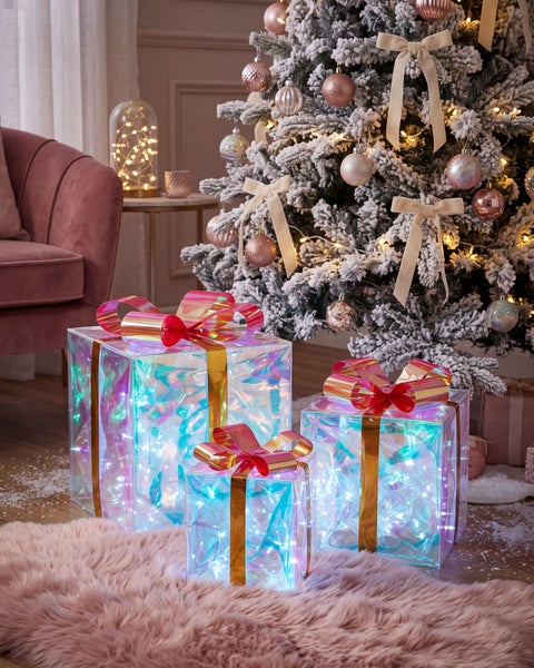 Drei beleuchtete quadratische Geschenkboxen mit irisierender Oberfläche und Schleifen unter einem verschneiten Weihnachtsbaum im Wohnzimmer.
