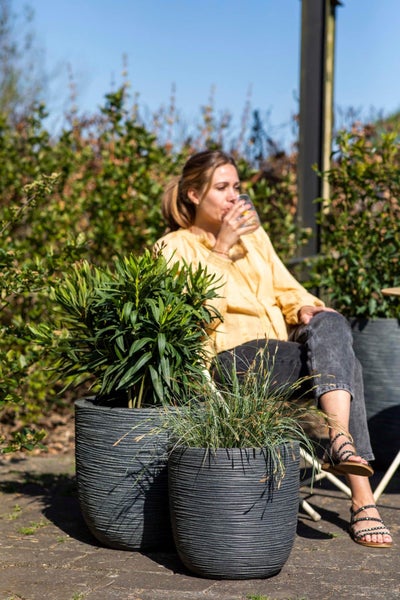 Frau sitzt in einem hellen Garten neben großen grauen Pflanzkübeln mit Rillenstruktur, bepflanzt mit Grünpflanzen und Ziergras.