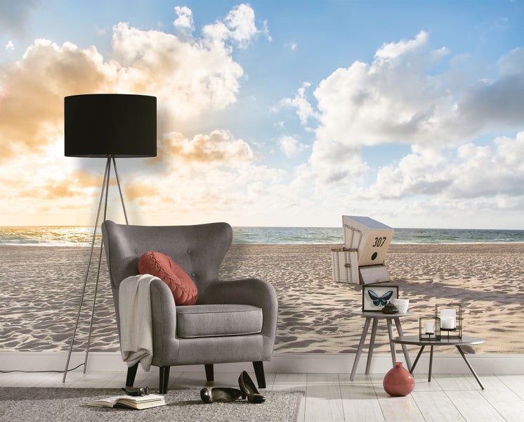 Fototapete mit Strandmotiv, Meer und Sonnenuntergang in einem Wohnzimmer mit grauem Sessel und schwarzer Stehlampe.