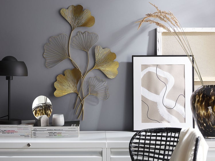 Dekoratives Interieur mit einer goldenen Wanddekoration in Blattform, einer Lampe, Kunstwerken und Wohnaccessoires auf einem weißen Tisch.