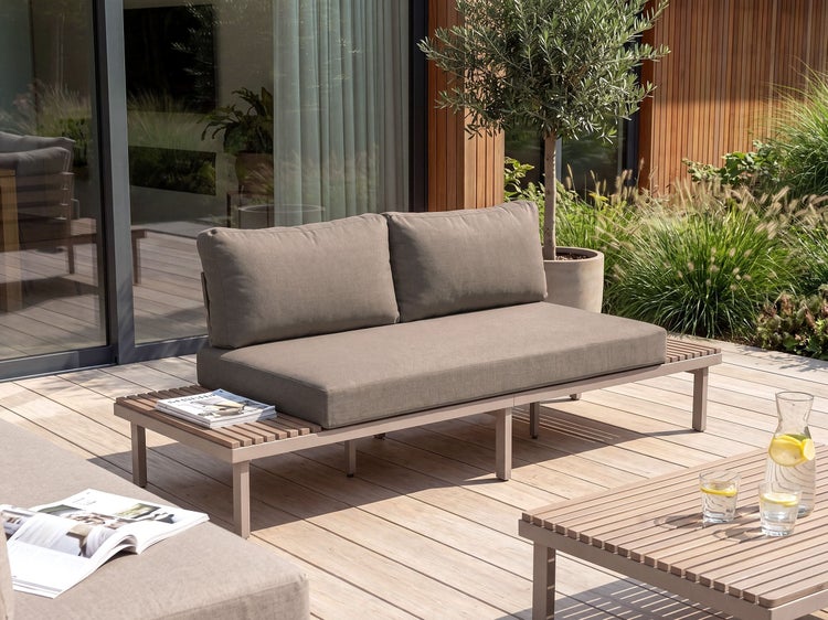 Modernes Gartensofa mit bequemen Polsterauflagen und integrierter Ablagefläche auf einer Holzterrasse vor einer modernen Hausfassade.