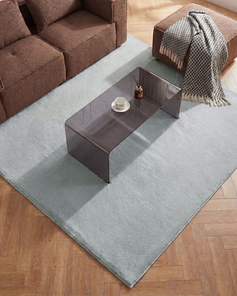 Hellblauer, flauschiger Hochflor-Teppich in einem modern eingerichteten Wohnzimmer mit braunem Sofa, Glastisch und Parkettboden.