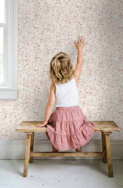 Vliestapete mit floralem Muster und Hasenmotiven in einem hellen Kinderzimmer, ein Kind sitzt auf einer Holzbank davor.
