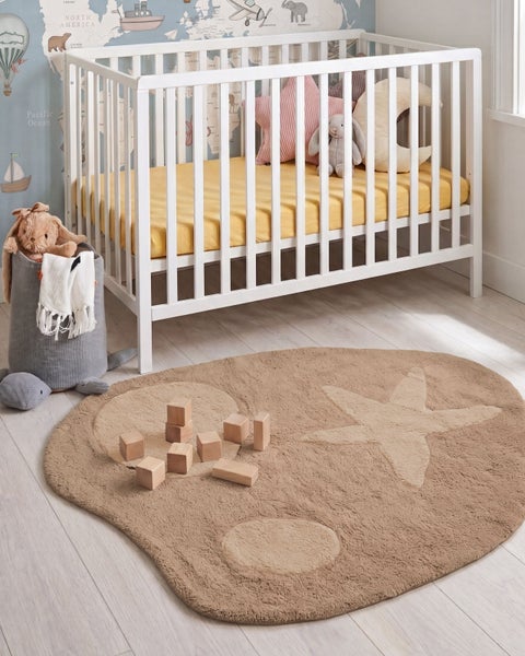 Ein Kinderzimmer mit einem weißen Gitterbett vor einer Weltkartentapete und einem runden, beigefarbenen Teppich mit Sternenmuster auf dem Boden.