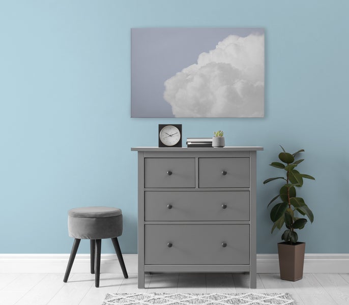 Graue Kommode mit drei Schubladen neben einem Hocker und einer Zimmerpflanze vor einer Wand mit Wolkenbild