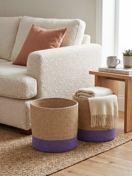Zwei geflochtene Aufbewahrungskörbe aus Jute mit lilafarbenem Akzent in einem hellen Wohnzimmer neben einem Sofa.