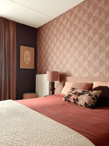 Schlafzimmer mit geometrisch gemusterter Tapete, Bett mit Kissen und Steppdecke, Tischleuchte und Wandbild in warmer Atmosphäre.