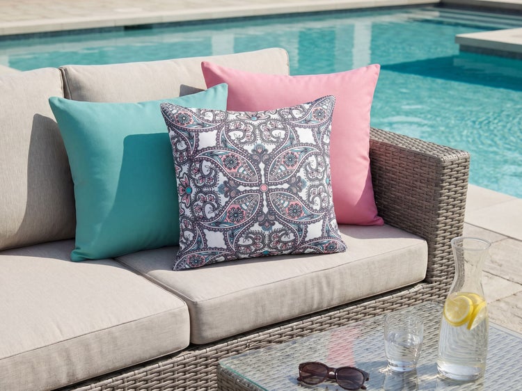 Dekorative Outdoor-Kissen mit Mandala-Muster und einfarbige Kissen auf einem grauen Lounge-Sofa aus Geflecht an einem Pool.