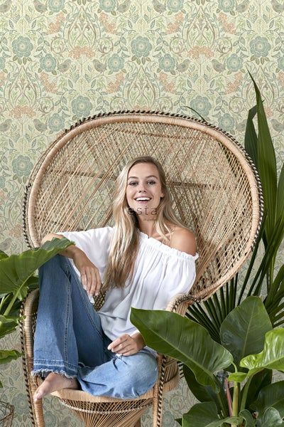 Frau sitzt in einem Rattansessel vor einer gemusterten Tapete und Pflanzen