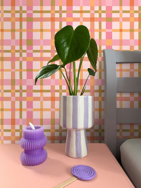 Vliestapete mit Karomuster in Rosa und Orange hinter einer Monstera Pflanze im gestreiften Übertopf und einer lila Kerze auf einem Nachttisch.
