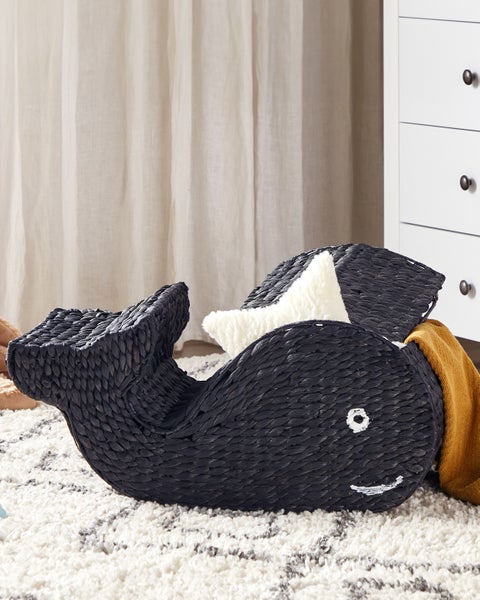 Geflochtener Aufbewahrungskorb in Walform aus dunklem Material mit Deckel, gefüllt mit Decke und Kissen auf einem hellen Teppich.