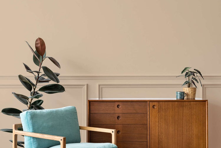 Hell eingerichtetes Wohnzimmer mit beigefarbener Wand mit Zierleisten, hölzernem Sideboard, hellblauem Sessel und Zimmerpflanzen.