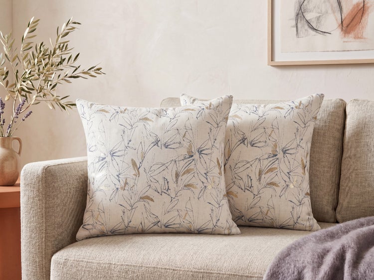 Zwei quadratische Deko-Kissen mit blauem und goldenem Blattmuster auf einem beigen Sofa in einem hellen Wohnzimmer.