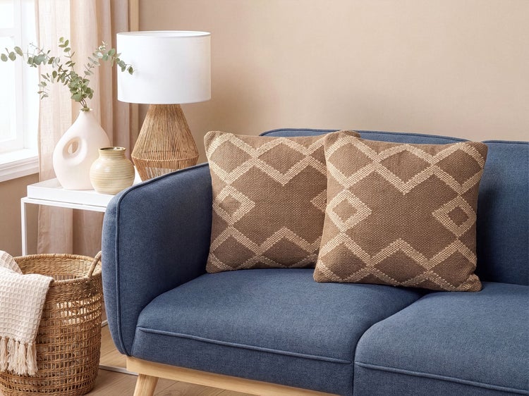 Zwei quadratische Dekokissen mit Rautenmuster auf einem blauen Sofa in einem wohnlich eingerichteten Raum mit Tischlampe und Korb.