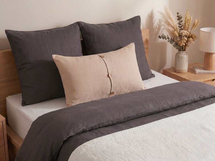 Modernes Schlafzimmer mit dunkelgrauer Bettwäsche, beigem Zierkissen mit Knöpfen und strukturierter weißer Decke auf einem Holzrahmen.