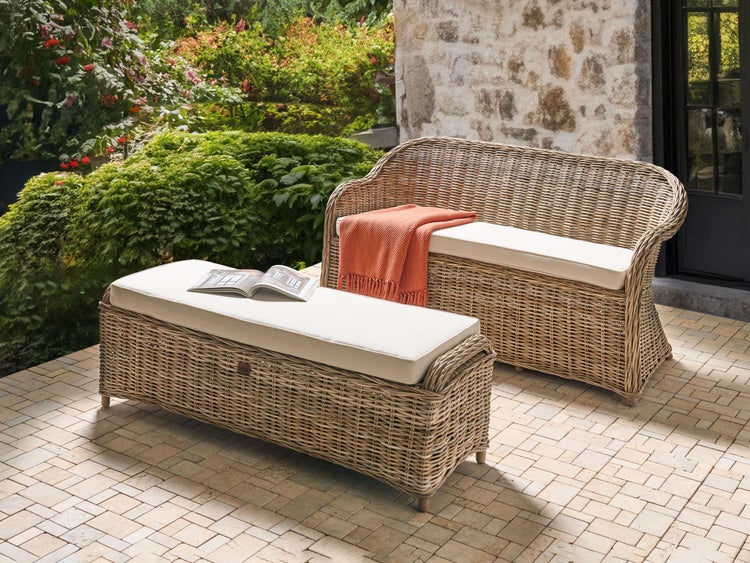 Gartenbank und Hocker aus Geflecht mit hellen Sitzkissen auf einer Terrasse vor einer Steinmauer in heller und organisierter Atmosphäre.