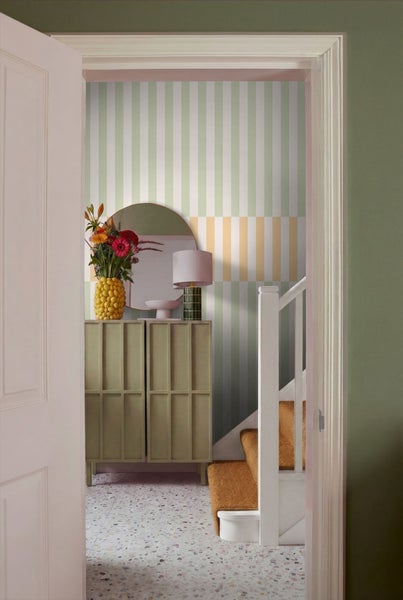 Blick durch eine offene Tür auf ein stilvolles Interieur mit Kommode, Spiegel, Lampe und Treppe