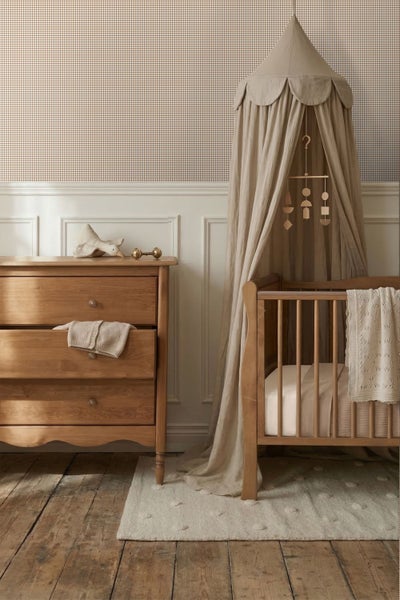 Hell eingerichtetes Kinderzimmer mit karierter Tapete, weißen Wandpaneelen, einer Holzkommode und einem Babybett mit Betthimmel.