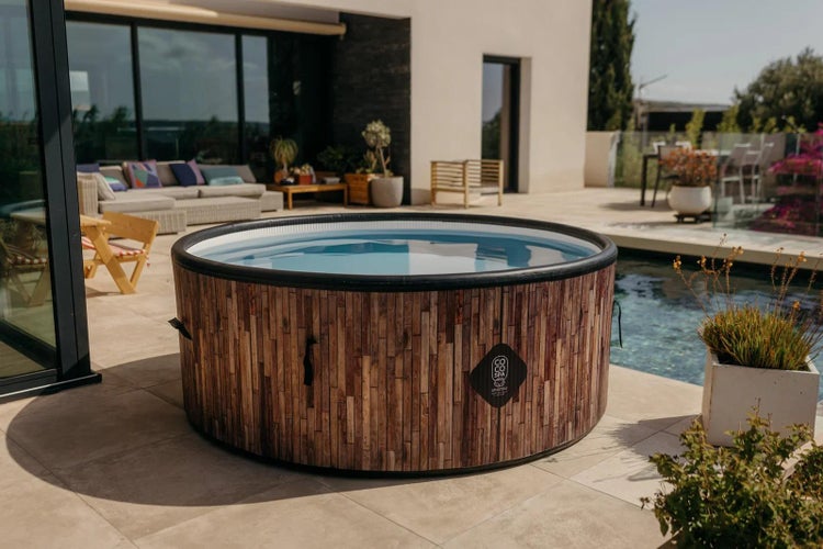 Runder aufblasbarer Whirlpool in Holzoptik mit CoCo Spa Logo auf einer hellen, modernen Terrasse neben einem Pool und Loungemöbeln.