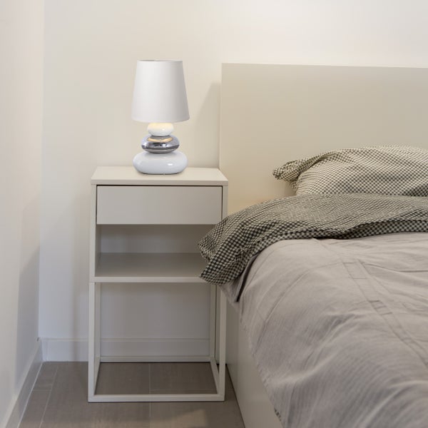 Nachttisch mit Schublade und darauf stehender Tischlampe neben einem Bett.