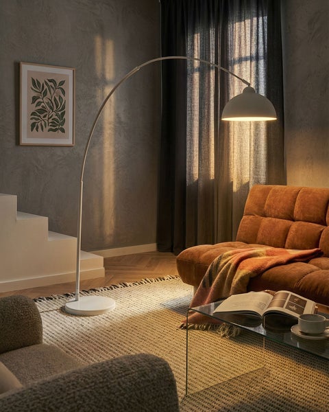 Moderne weiße Bogenstehleuchte in einem gemütlichen Wohnzimmer mit braunem Sofa, Glastisch und strukturiertem Teppich vor grauer Wand.