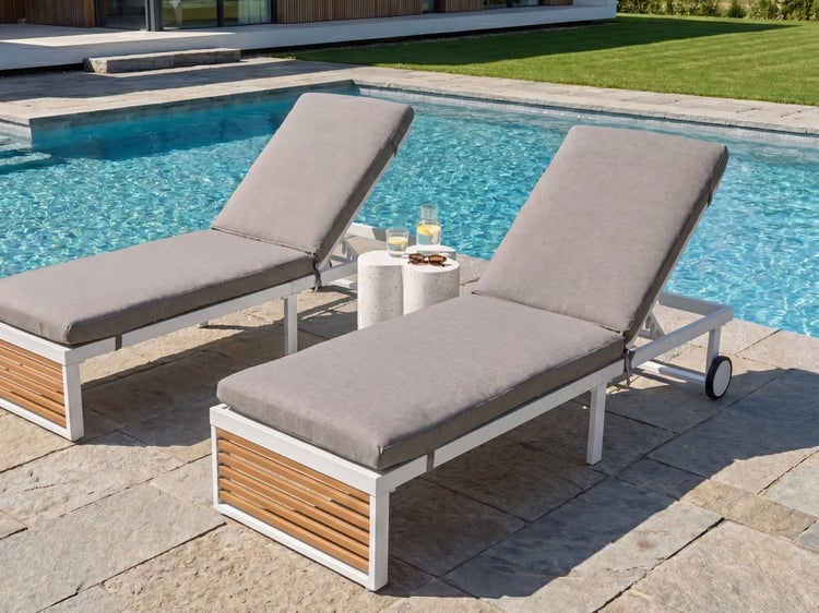 Zwei moderne Sonnenliegen mit grauem Polster und Holzdekor auf einer Steinterrasse neben einem Swimmingpool.
