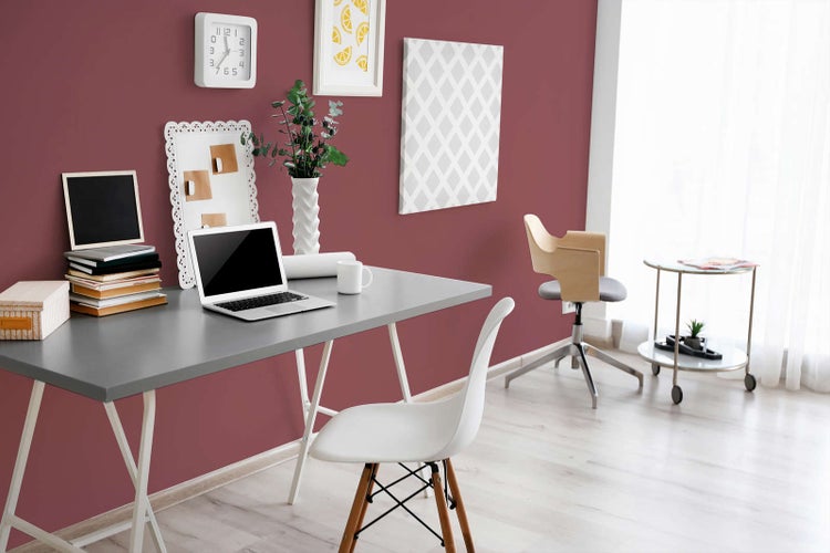 Modernes Arbeitszimmer mit beerenfarbener Wandfarbe, einem grauen Schreibtisch mit Laptop, einem weißen Stuhl und hellem Boden in Holzoptik.