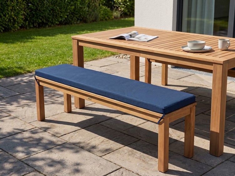 Gartenbank aus Holz mit einem aufliegenden Sitzpolster auf einer Terrasse mit Gartentisch im Hintergrund.