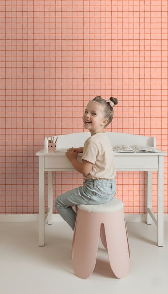 Mädchen sitzt an einem Schreibtisch auf einem Hocker vor einer Wand mit einem quadratischen Muster.