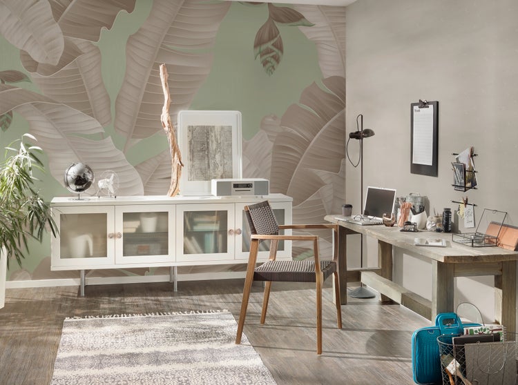 Hell gestaltetes Arbeitszimmer mit tropischer Fototapete mit großen Bananenblättern, weißem Sideboard, Holzschreibtisch und geflochtenem Stuhl.