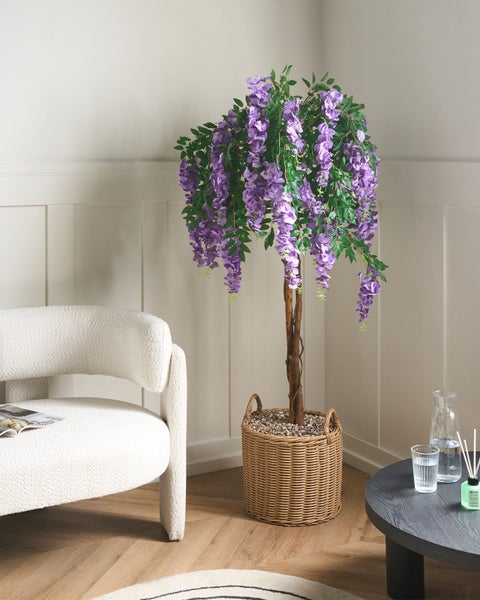 Künstlicher Blauregen mit lila Blüten in einem Weidenkorb, dekoriert in einem hellen Wohnzimmer mit weißem Sessel.