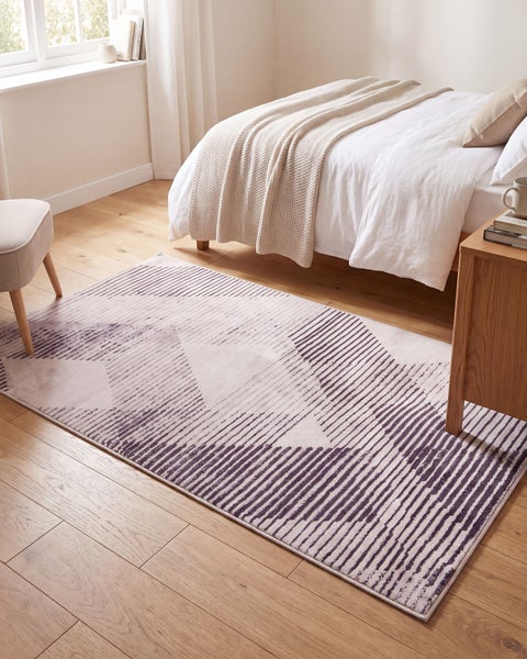 Moderner Teppich mit geometrischem Streifenmuster vor einem Bett im hellen, wohnlichen Schlafzimmer mit Holzboden.