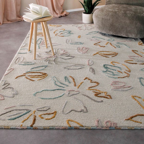 Hell gestalteter Wohnraum mit floral gemustertem Teppich, einem Holzhocker, einer Pflanze und einem grauen Samtpouf.