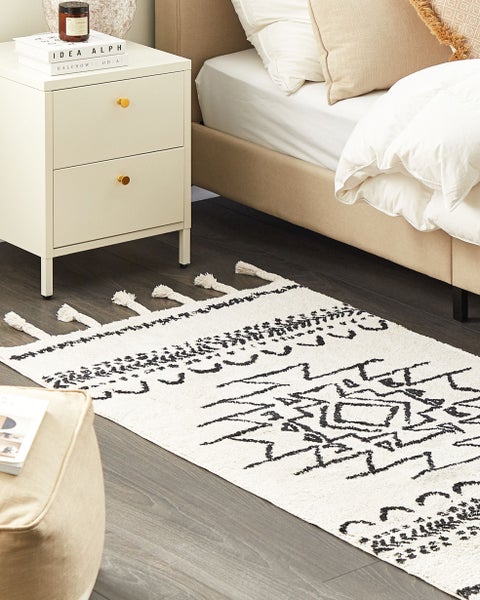 Hell gestaltetes Schlafzimmer mit einem rechteckigen Teppich mit geometrischem Muster und Quasten, einem Bett und einem Nachttisch.