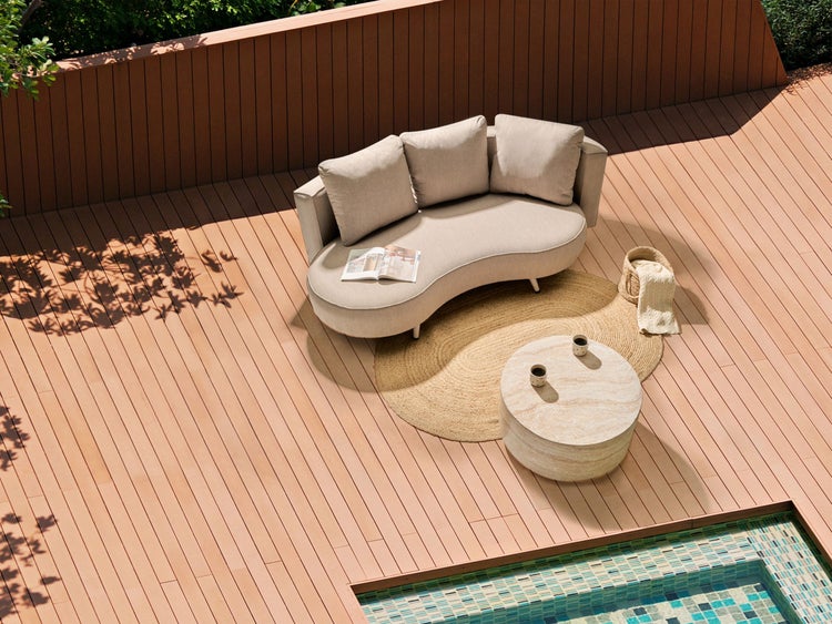 Geschwungenes Loungesofa mit Kissen, runder Beistelltisch in Steinoptik und Juteteppich auf einer Holzterrasse am Pool.