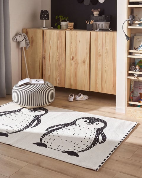 Kinderspielzimmer mit Teppich mit Pinguinmuster, gestreiftem Sitzhocker, Holzkommode und einem Bücherregal aus hellem Kiefernholz.