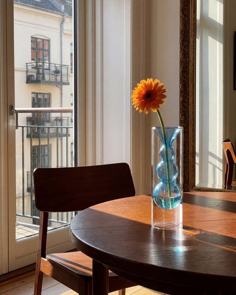 Eine zylindrische Glasvase mit blauem Innenteil und einer orangefarbenen Gerbera auf einem runden Holztisch in einem hellen Raum.